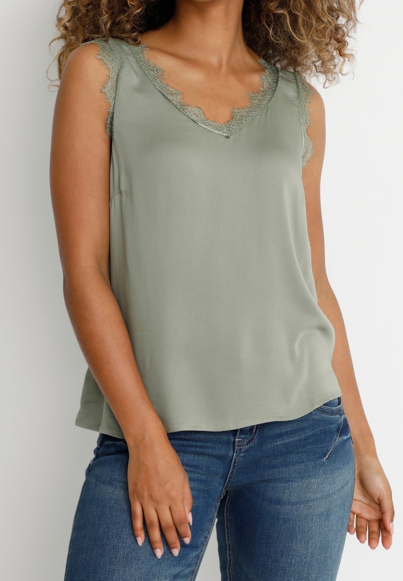 Femme portant un haut sans manches vert sauge avec bordure en dentelle et un jean en denim bleu, debout devant un fond clair uni.
