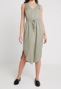 Lekka oliwkowa sukienka bez rękawów z dekoltem w serek, elastyczną talią i rozcięciami po bokach. Gładka tkanina i luźna, rozkloszowana sylwetka. Noszona z czarnymi balerinami.