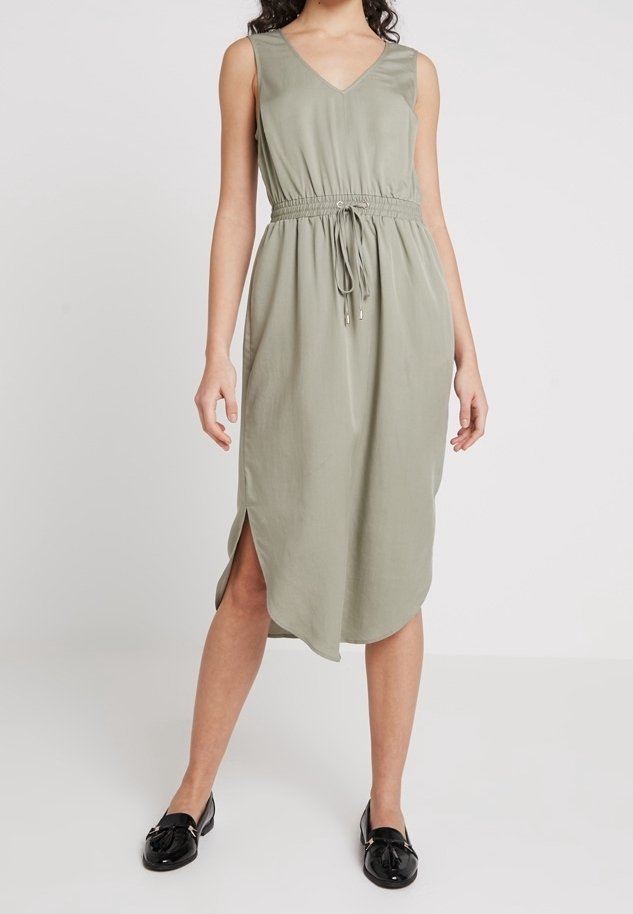 Lekka oliwkowa sukienka bez rękawów z dekoltem w serek, elastyczną talią i rozcięciami po bokach. Gładka tkanina i luźna, rozkloszowana sylwetka. Noszona z czarnymi balerinami.