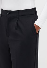 Black trousers with a tailored fit, featuring front pleats, side pockets, and a button closure at the waistband. Smooth fabric texture.