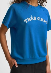 Mėlynas trumpa rankovėmis džemperis su raštuotu apykaklės iškirpimu, priekyje centre esantis baltas užrašas "TRÈS CHIC", pagamintas iš lygaus audinio.
