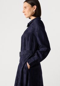 Navyblauwe jurk met een bloemenpatroon, lange mouwen, een ceintuur in de taille en zakken. De stof heeft een gladde textuur en een lichte glans.