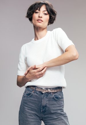 Suéter blanco de punto de manga corta con patrón texturizado, combinado con jeans de cintura alta de color azul deslavado y un cinturón con tachuelas.