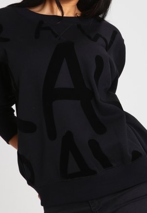 Femme portant un sweat-shirt noir avec de grandes lettres abstraites noires imprimées sur le devant, assorti à un pantalon noir.