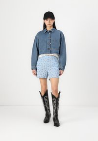 Τζάκετ από τζιν κομμένο με κούμπωμα, ανοιχτό μπλε σορτς με σχέδιο, ασημένια ζώνη και μαύρες καουμπόικες μπότες μέχρι το γόνατο με λευκές κεντητές λεπτομέρειες.