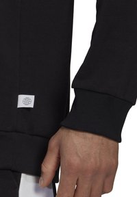 Sweatshirt noir à finition texturée, doté de poignets côtelés et d'un petit patch logo blanc près de l'ourlet. Tissu lisse, design décontracté.