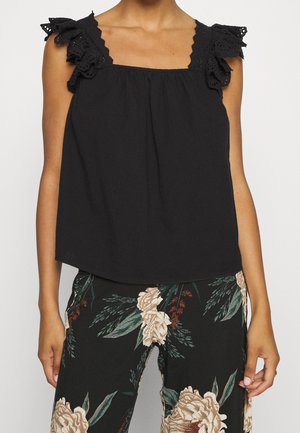 Woman wearing a sleeveless black top with ruffled lace straps and black pants with large beige floral print.