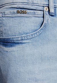 Vaqueros de mezclilla azul claro con un bolsillo frontal, acentuados por una etiqueta de logo "BOSS" en plata. Presentan costuras clásicas y una textura suave.