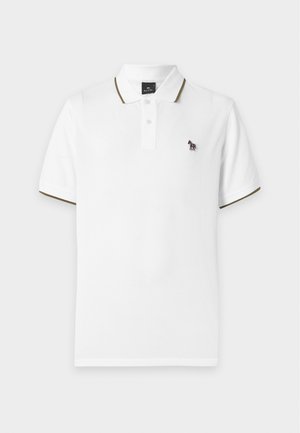 Jeune homme debout portant un polo blanc, un pantalon beige et des mocassins jaunes sur fond blanc uni.