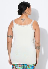 Top de tirantes color crema claro con encaje en el escote y los tirantes; diseño ajustado con una textura suave. Se lleva con pantalones de patrón colorido.