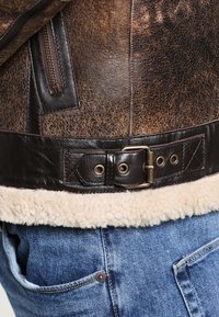 Veste en cuir marron avec doublure en shearling à l'ourlet. Comprend une boucle en métal et une poche zippée sur le côté, avec une surface texturée.