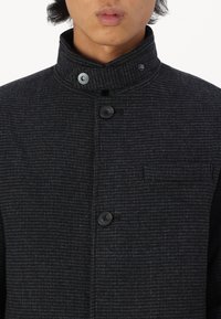 Svart teksturert jakke med høy krage, som har to knapper og en brystlomme. Stoffet har et subtilt rutemønster.