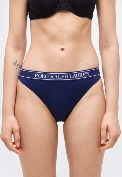 Γυναίκα που φοράει μπλε σκούρο ψηλόμεσο εσώρουχο με τη ζώνη μέσης "Polo Ralph Lauren" και μαύρο σουτιέν, εμφανίζοντας τον κορμό και το πάνω μέρος των μηρών.