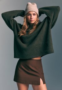 Pull en maille vert foncé oversize à manches longues, associé à une mini jupe marron avec une fente sur le côté, coiffé d'un bonnet beige clair.