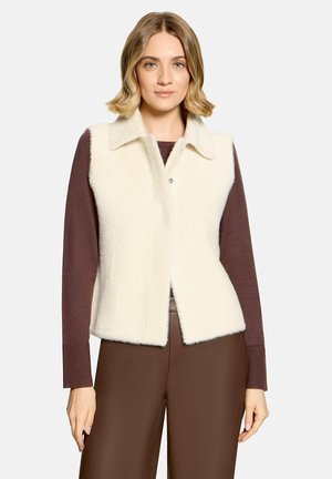 Kvinde med kort blondt hår iført en hvid fluffy vest over en brun langærmet top og brune bukser, stående mod en ensfarvet baggrund.