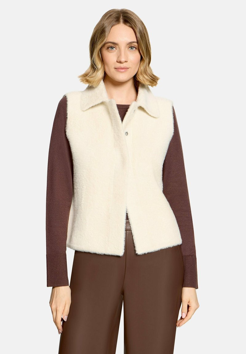Femme aux cheveux blonds courts portant une veste blanche duveteuse par-dessus un top marron à manches longues et un pantalon marron, debout devant un fond uni.