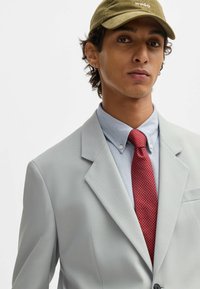 Light gray suit with textured fabric, blue button-down shirt, and a red patterned tie. A green cap is worn over curly hair.