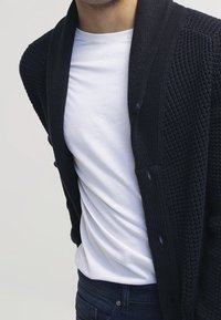 Mörkblå stickad kofta med sjalkrage, oversized passform, med stora knappar och texturerat material, som bärs över en vit t-shirt.