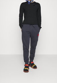 Haut noir à manches longues avec un accent diagonal, pantalon de jogging gris avec une poche latérale, et sandales colorées à brides.