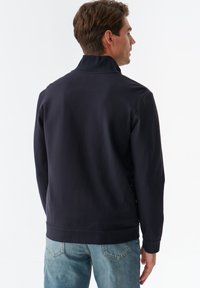 Marineblå sweatshirt med høj krave, glat tekstur og tætsiddende manchetter. Parret med lyseblå jeans med baglommer.