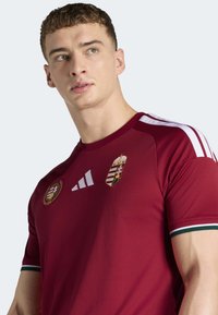 Rød atletisk trøje med hvide striber på skuldrene, med Ungarns nationalemblem og logoer. Blødt stof med en tætsiddende pasform.