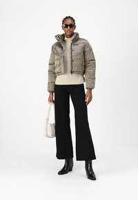 Giacca puffer verde oliva tagliata, maglione crema a coste, pantaloni neri a zampa, stivali neri con tacco e una borsa crema con tracolla logo.