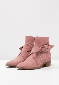 Bottes de cheville en suède rose avec un détail de nœud enroulé, bout pointu et un petit talon en bois. Fermeture à glissière sur le côté.
