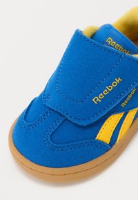 Zapatilla de ante azul con acentos amarillos y tira de velcro. Punta redondeada y suela de goma. Marca Reebok en amarillo en el costado y la tira.