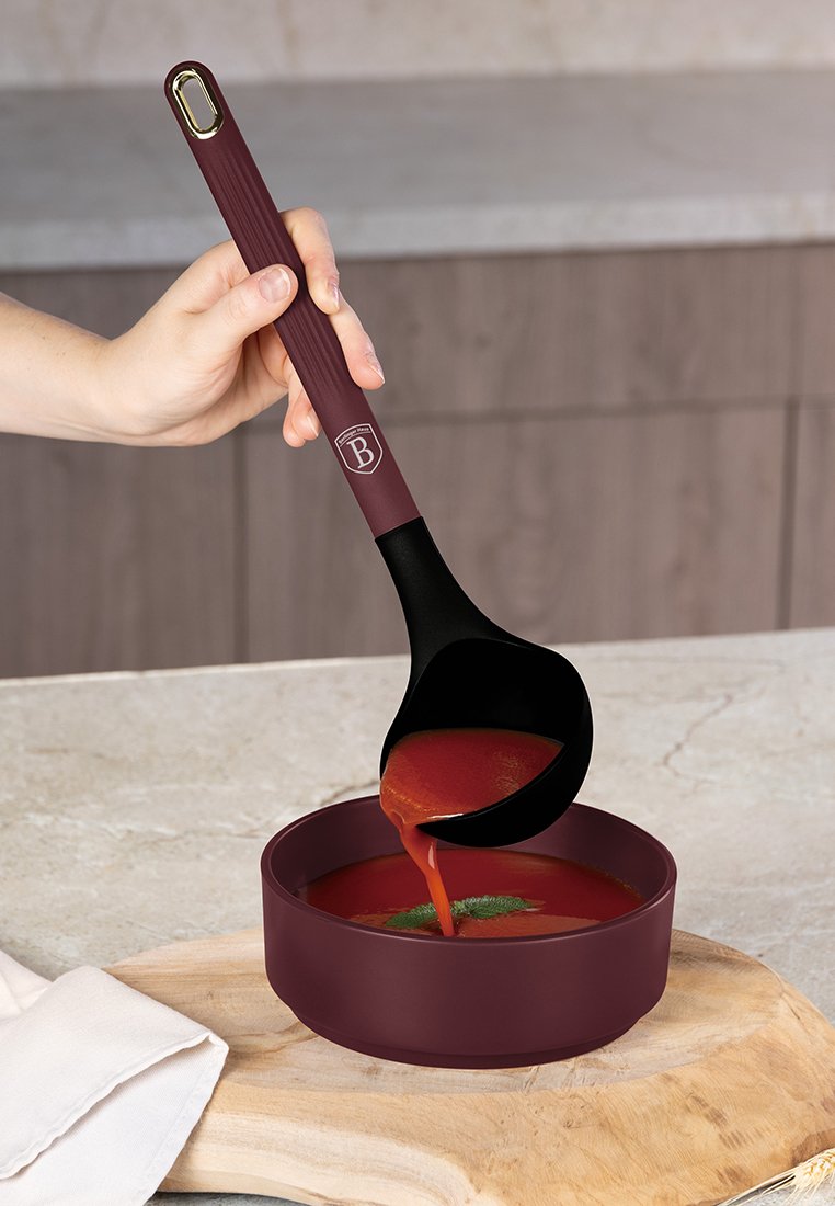 Hand, die eine burgunderfarbene Schöpfkelle hält und Tomatensuppe mit Garnitur in eine passende Schüssel auf einem Holzbrett und einer Marmorarbeitsplatte gießt.