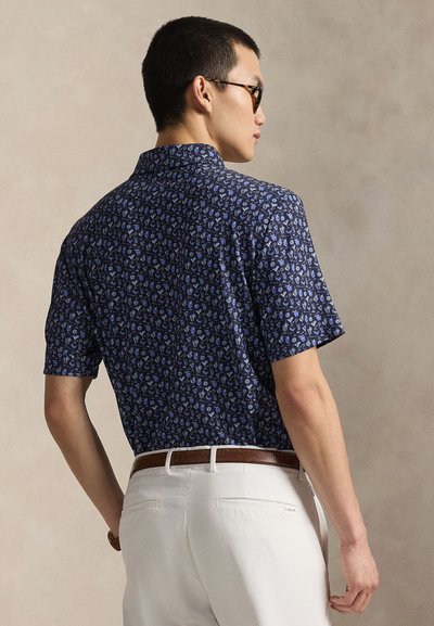 Κοντομάνικο πουκάμισο σε ναυτικό μπλε με μικρό, επαναλαμβανόμενο ανοιχτό μπλε μοτίβο, σχεδιασμό με γιακά και ίσια βάση, συνδυασμένο με ανοιχτόχρωμο παντελόνι.