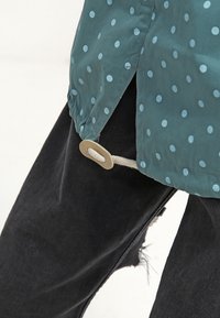 Turkusowa kurtka z niebieskimi groszkami, wyposażona w boczny rozcięcie i okrągły beżowy guzik jako akcent. Czarne, postarzane dżinsy z widocznymi przetartymi brzegami.