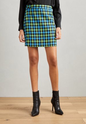 Jupe à carreaux courte en bleu, vert et noir avec un design ajusté. Portée avec des bottines noires, mettant en valeur un tissu texturé et des lignes nettes.