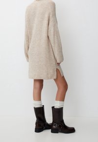 Beżowa dzianinowa sukienka z dużymi rękawami i bocznymi rozcięciami, zestawiona z błyszczącymi brązowymi kozakami za kolano i kremowymi skarpetkami.