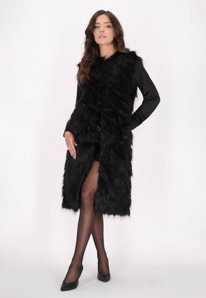 Donna che indossa un lungo gilet nero in pelliccia sintetica sopra un vestito nero, collant neri trasparenti e tacchi neri, in piedi con una mano sui fianchi.