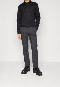 Chemise noire à manches longues avec boutons, associée à un pantalon à carreaux gris. Chaussures : bottes noires à lacets avec semelles épaisses et coutures jaunes.
