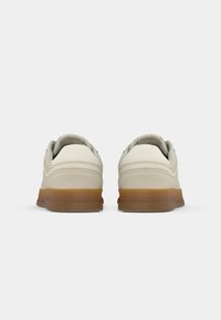 Paire de baskets blanc cassé avec semelles marron clair vues de dos sur un fond gris clair uni.