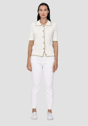 Femme debout face à l’avant, portant un haut blanc à manches courtes boutonné avec bordure beige, pantalon blanc, et baskets blanches sur un fond uni.