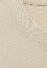 Materialul din bumbac bej pentru tricou are un guler rotund ribatat și o textură netedă. Detaliile de cusătură de-a lungul gulerului adaugă un accent subtil.