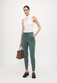 Groene rekbare broek met zijzakken, voorzien van een taillekoord en een gladde textuur, gecombineerd met een aansluitend wit topje en een bruine handtas.