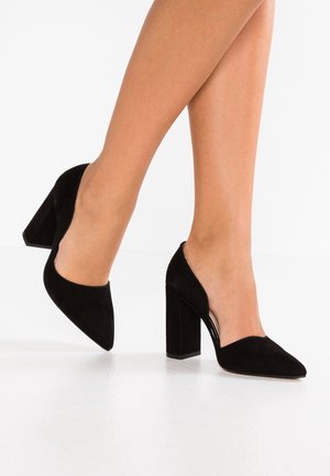Jambes portant des chaussures à talons blocs en suède noir à bout pointu, debout sur un sol blanc devant un fond blanc.