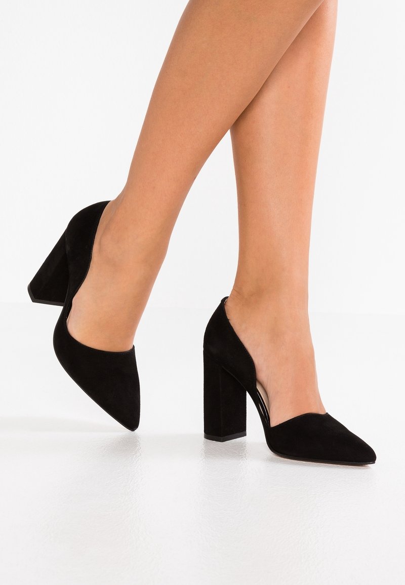 Jambes portant des chaussures à talons blocs en suède noir à bout pointu, debout sur un sol blanc devant un fond blanc.