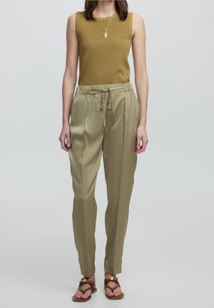 Kvinde iført brun ærmeløs top, beige højtaljede bukser med snøring, guldfarvet vedhængs halskæde og brune sandaler med gulddetaljer.