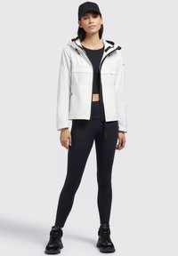 Veste imperméable blanche avec capuche, poches avant et accents noirs contrastants. Portée par-dessus un crop top noir et des leggings noirs. Baskets noires.