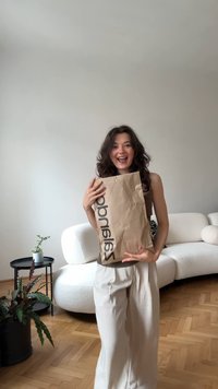 Brązowa torba papierowa z czarnym napisem "salando", trzymana przed osobą w beżowych szerokich spodniach w minimalistycznym pomieszczeniu.