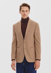 W kratkę blazer w brązowo-beżowym materiale, zapinany na jeden guzik, z klapami, noszony na ciemnym golfie, w zestawieniu z niebieskimi dżinsami.