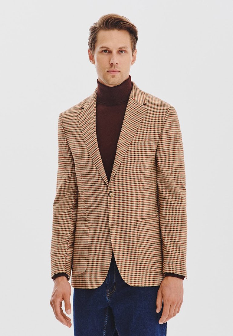 W kratkę blazer w brązowo-beżowym materiale, zapinany na jeden guzik, z klapami, noszony na ciemnym golfie, w zestawieniu z niebieskimi dżinsami.