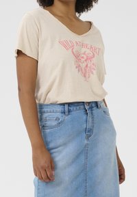 Béžové tričko s krátkým rukávem a kulatým výstřihem, s růžovým grafickým designem obsahujícím květinové a býčí motivy. V kombinaci se světle modrou džínovou sukní.