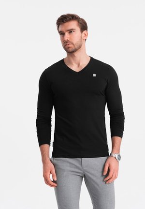Camisa negra de manga larga y cuello en V, hecha de una tela lisa y ligera. Presenta un pequeño logo en el pecho. Combinada con pantalones gris claros.
