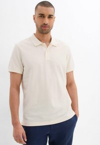 Polo a maniche corte di un leggero beige realizzato in tessuto morbido. Presenta un colletto classico e una patta con due bottoni. Design semplice e pulito.