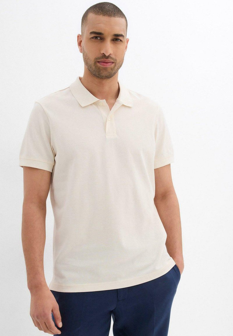 Polo a maniche corte di un leggero beige realizzato in tessuto morbido. Presenta un colletto classico e una patta con due bottoni. Design semplice e pulito.